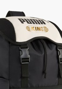 Zaino con corpo nero, flap bianca con "PUMA" e "KING" in oro, dettagli in rete e accessori neri. Cinghie regolabili incluse.