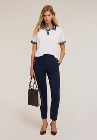 Tricou polo alb cu guler în dungi albastre și roșii, asortat cu pantaloni slim-fit negri cu detalii din butoane aurii. Poartă o poșetă multicoloră.