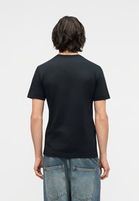 Άνδρας με μεσαίου μήκους καστανά μαλλιά, φορώντας ένα απλό μαύρο κοντομάνικο μπλουζάκι και ανοιχτό μπλε τζιν, κοιτάζοντας από πίσω.