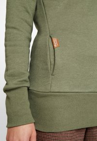 Sudadera verde con dobladillo y puños acanalados, bolsillo lateral con etiqueta de cuero marrón. La tela parece suave y texturizada.