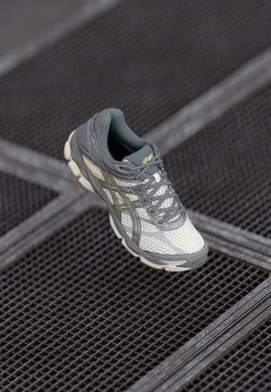 Grauer und weißer Asics Laufschuh mit gelben Akzenten und Schnürsenkeln, der auf einer strukturierten Metallgitteroberfläche ruht.