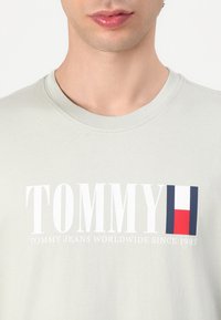 Light gray cotton t-shirt featuring "TOMMY" in bold white letters and a flag emblem with red, white, and blue accents.