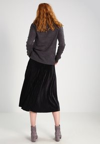 Grauer Strickpullover, schwarze plissierte Midirock, braune Stiefeletten. Fokus auf die Struktur und die vertikalen Linien des Rocks. Schlichte, elegante Silhouetten.