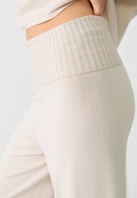 Ingrandimento di una persona che indossa pantaloni a maglia beige a vita alta e a coste, con una fascia larga in vita, che mostra il lato e l'area dei fianchi.