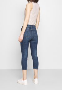 Jeans de denim azul oscuro con cintura de media altura, corte ajustado y largo recortado, combinados con una camiseta sin mangas de canalé beige claro y zapatillas blancas.