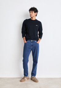 Herrtröja i marinblått med rund hals och logotyp, ihopparad med löst sittande blå jeans och ljusbruna sneakers. Minimalistisk design, avslappnad stil.