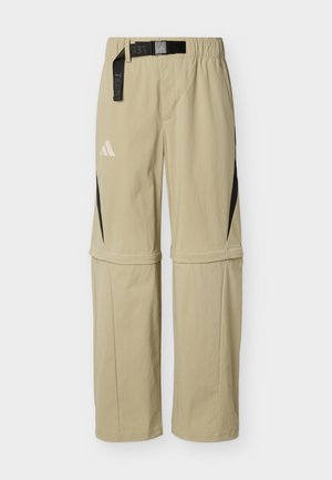 Gaismas bēša krāsas pārveidojamās āra bikses ar melnu jostu un sānu rāvējslēdzēju paneļiem, uz kreisās augšstilba daļas redzams mazs balts logo.