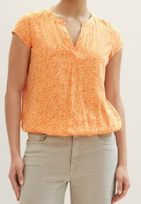 Blouse orange à manches courtes avec un motif floral, col en V, tissu léger et coupe décontractée, portée avec un jean de couleur claire.
