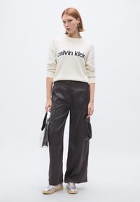 Krémový pletený sveter s nápisom "calvin klein", v kombinácii s čiernymi širokými nohavicami. Strieborné tenisky a metalická kabelka dopĺňajú vzhľad.