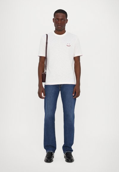 T-shirt blanche avec des accents rouges et un design de visage souriant, assortie à un jean bleu et des chaussures noires. Inclus un sac en bandoulière marron.