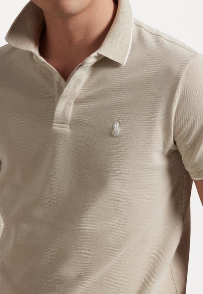 Vaaže paita beige värviga, millel on tekstuurne kanga struktuur, kahest nööbist plaan ja tõstetud krae. Vasakul rinnal on väike valge logo.