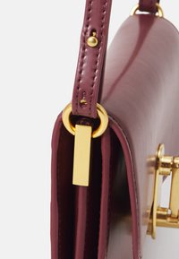 Borsa in pelle bordeaux con una texture liscia, dettagli in hardware dorato e un'etichetta rettangolare distintiva attaccata alla tracolla.