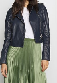 Giacca da motociclista in pelle blu navale con tasche con zip, hardware argentato e design aderente, abbinata a una gonna plissettata in raso verde.