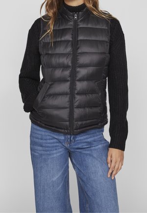 Schwarze, gepolsterte Weste mit hohem Kragen und Reißverschluss, ausgestattet mit gestrickten schwarzen Ärmeln und vorderen Taschen, kombiniert mit blauen weit geschnittenen Jeans.