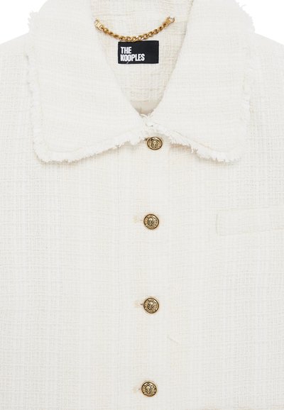 Veste blanche texturée avec un large col effiloché, des détails en chaîne dorée, et quatre boutons décoratifs dorés à l'avant.
