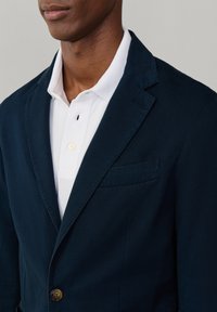 Homme portant un blazer bleu marine par-dessus une chemise blanche à col, visible du cou jusqu'au milieu du torse sur un fond uni.