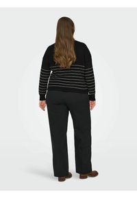 Femme aux longs cheveux bruns portant un pull à rayures noires, un pantalon noir et des chaussures marron, vue de dos sur un fond clair uni.
