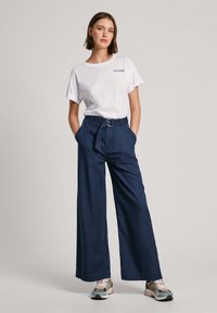 Witte katoenen t-shirt met opgerolde korte mouwen en marineblauwe wijde broek met een riem met een ronde zilveren gesp, gecombineerd met kleurrijke sneakers.