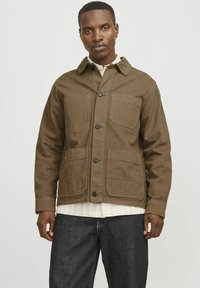 Chaqueta de lona marrón con cuatro bolsillos, cierre de botón y cuello tipo solapa. Lleva puesta una camisa blanca a rayas y jeans oscuros.