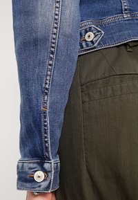 Kurtka jeansowa w ciemnoniebieskim kolorze, z kontrastującymi szwami i detalami z zatrzaskami, zestawiona z oliwkowozielonym materiałem pod spodem.