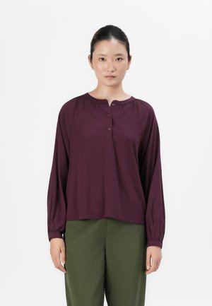 Femme aux cheveux bruns attachés, portant un chemisier prune à manches longues avec des boutons et un pantalon vert olive, se tenant devant un fond blanc.