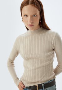 Beżowy, prążkowany top z długim rękawem i wysokim kołnierzem, wykonany z miękkiego materiału. Charakteryzuje się dopasowanym krojem i subtelnymi pionowymi paskami.