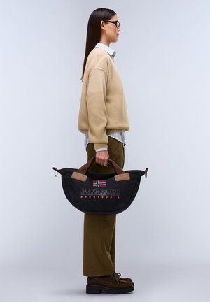 Czarna torebka z tkaniny ze brązowymi akcentami, o zaokrąglonym kształcie, z logo i detalem w postaci flagi, niesiona przez osobę w beżowym swetrze i oliwkowo-zielonych spodniach.