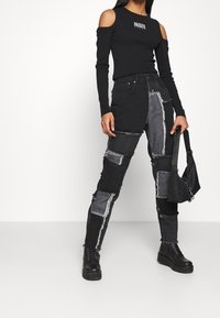 Top nero off-shoulder a maniche lunghe con logo "RAGGED", abbinato a jeans in denim nero a patchwork con sfumature diverse e bordi sfilacciati. Stivali neri e borsa.