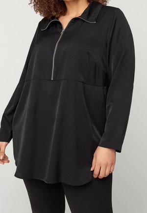 Femme portant un haut noir ample à manches longues avec une fermeture éclair devant et un motif texturé subtil, associé à un pantalon noir.
