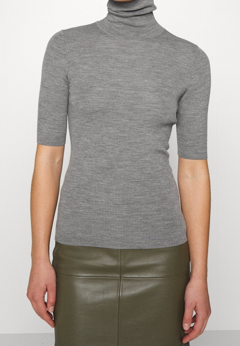 Personne portant un pull gris côtelé à manches courtes et col roulé, ainsi qu'une jupe en cuir vert olive foncé, debout sur un fond blanc uni.
