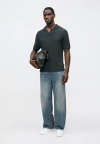 Σκούρο γκρι πόλο πουκάμισο με κοντά μανίκια και υφασμάτινη υφή. Συνδυάζεται με φαρδιά τζιν παντελόνια ξεθωριασμένου μπλε χρώματος και λευκά αθλητικά παπούτσια. Κρατάει μαύρη τσάντα.