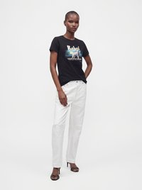 Γυναίκα που φοράει μαύρο γραφικό t-shirt "Rebel", λευκά τζιν με επιθέματα σε σχήμα καρδιάς στα γόνατα και μαύρα σανδάλια με τακούνι, στεκόμενη μπροστά σε απλό φόντο.