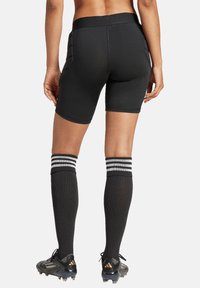 Schwarze Sportshorts mit mittelhoher Taille, seitlichen Einsätzen und glattem Material, kombiniert mit knielangen schwarzen Socken mit weißen Streifen.