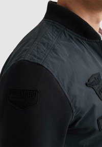 Nærbillede af en person iført en sort og mørkegrå bomberjakke med tekstureret ribbet krave og påsyede lapper på skulder og overarm.