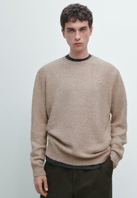 Maglione lavorato a maglia di un marrone chiaro con colletto rotondo, bordi a coste e una texture morbida, indossato sopra una camicia scura, abbinato a pantaloni scuri.
