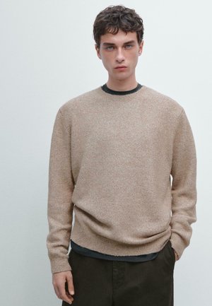 Maglione lavorato a maglia di un marrone chiaro con colletto rotondo, bordi a coste e una texture morbida, indossato sopra una camicia scura, abbinato a pantaloni scuri.