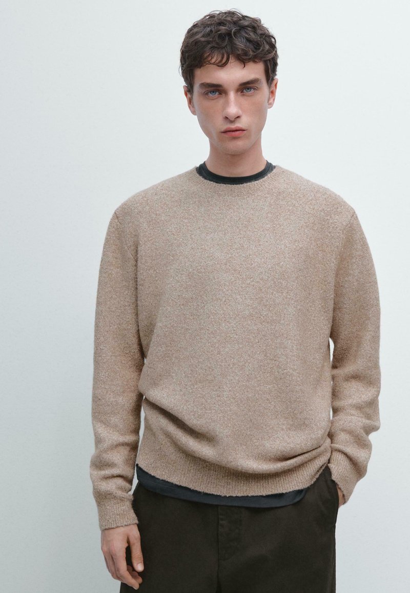 Maglione lavorato a maglia di un marrone chiaro con colletto rotondo, bordi a coste e una texture morbida, indossato sopra una camicia scura, abbinato a pantaloni scuri.