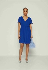 Ženska v rahlo dolgi kraljevo modri obleki z letečimi rokavi, srebrnih čevljih, drži svetlo modro puhasto torbico pred enobarvno ozadje.