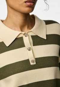 Zbliżenie na pasiastą zielono-beżową dzianinową koszulkę polo z trzema ozdobnymi złotymi guzikami i kołnierzykiem, noszoną przez osobę.