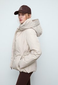 Giacca imbottita beige con colletto alto e cappuccio grande. Presenta una vita con cintura, chiusura con zip e automatici, e tasche laterali. Texture lucida.