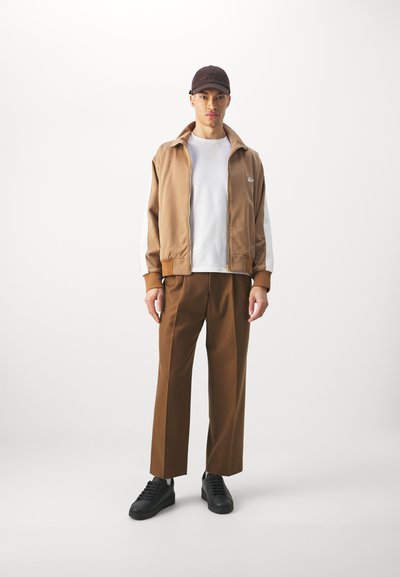 Giacca marrone con zip e maniche bianche, scarpe da ginnastica nere, pantaloni marroni e un cappello scuro. Modello in piedi contro uno sfondo bianco semplice.
