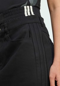 Černé kalhoty s texturovanou tkaninou, s elastickým páskem a třemi bílými pruhy. Viditelné postranní kapsy a podrobnosti s vertikálním švem.