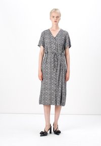 Vestido de padrão floral em preto e branco com mangas curtas, decote em V e cintura com cinto. A saia cai abaixo do joelho. Sapatos de salto alto pretos.