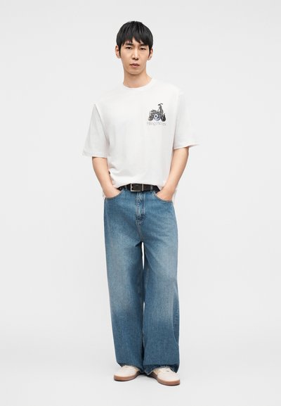 Λευκό κοντομάνικο μπλουζάκι με γραφικό σχέδιο. Χαλαρή εφαρμογή μπλε τζιν παντελόνι, στυλ με φαρδιά πόδια. Κρεμ αθλητικά παπούτσια με ανοιχτό καφέ σόλα. Μαύρη ζώνη.