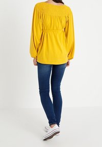 Gul smock-stil topp med smockdetaljer vid axlarna, kombinerad med mörkblå skinny jeans och vita sneakers.