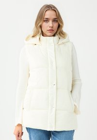 Gepolsterte, cremefarbene Weste mit Kapuze, die Druckknöpfe und seitliche Taschen hat. Das Außenmaterial ist glatt mit leichtem Glanz und einem leichten Textur.