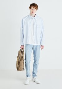 Camisa de botones de rayas azul claro sobre una camiseta blanca, jeans azul claro, zapatos blancos, sosteniendo una mochila de tela marrón.