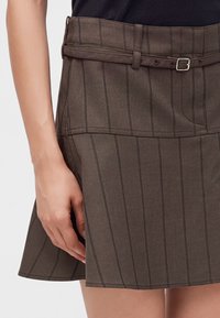 Femme portant une jupe marron à fines rayures avec une ceinture fine assortie et un top noir, main reposant sur le côté