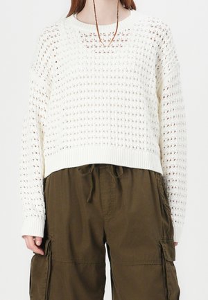 Osoba ubrana w biały, ażurowy sweter z długimi rękawami oraz brązowe spodnie bojówki z kieszeniami i ściągaczem w talii.