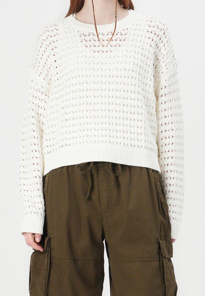 Person wearing a white, open-knit sweater with long sleeves and brown cargo pants with pockets and a drawstring waist.